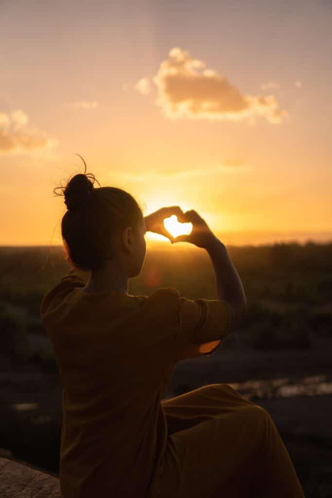 Jente som lager et hjerte med hendene foran en solnedgang