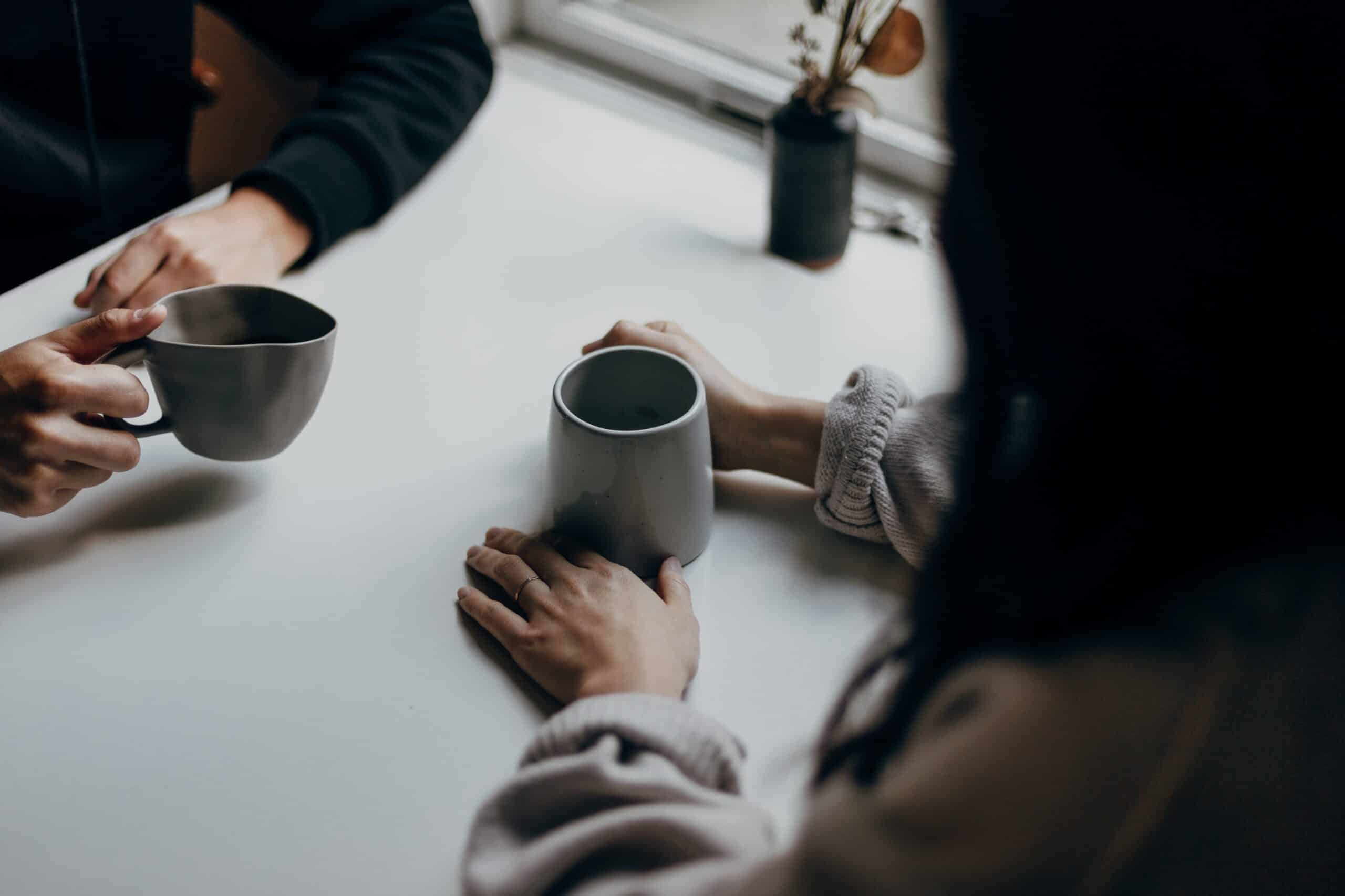 Illustrasjonsbilde som viser to personer som sitter ved et bord med en kaffekopp. Begge har hendene på bordet.