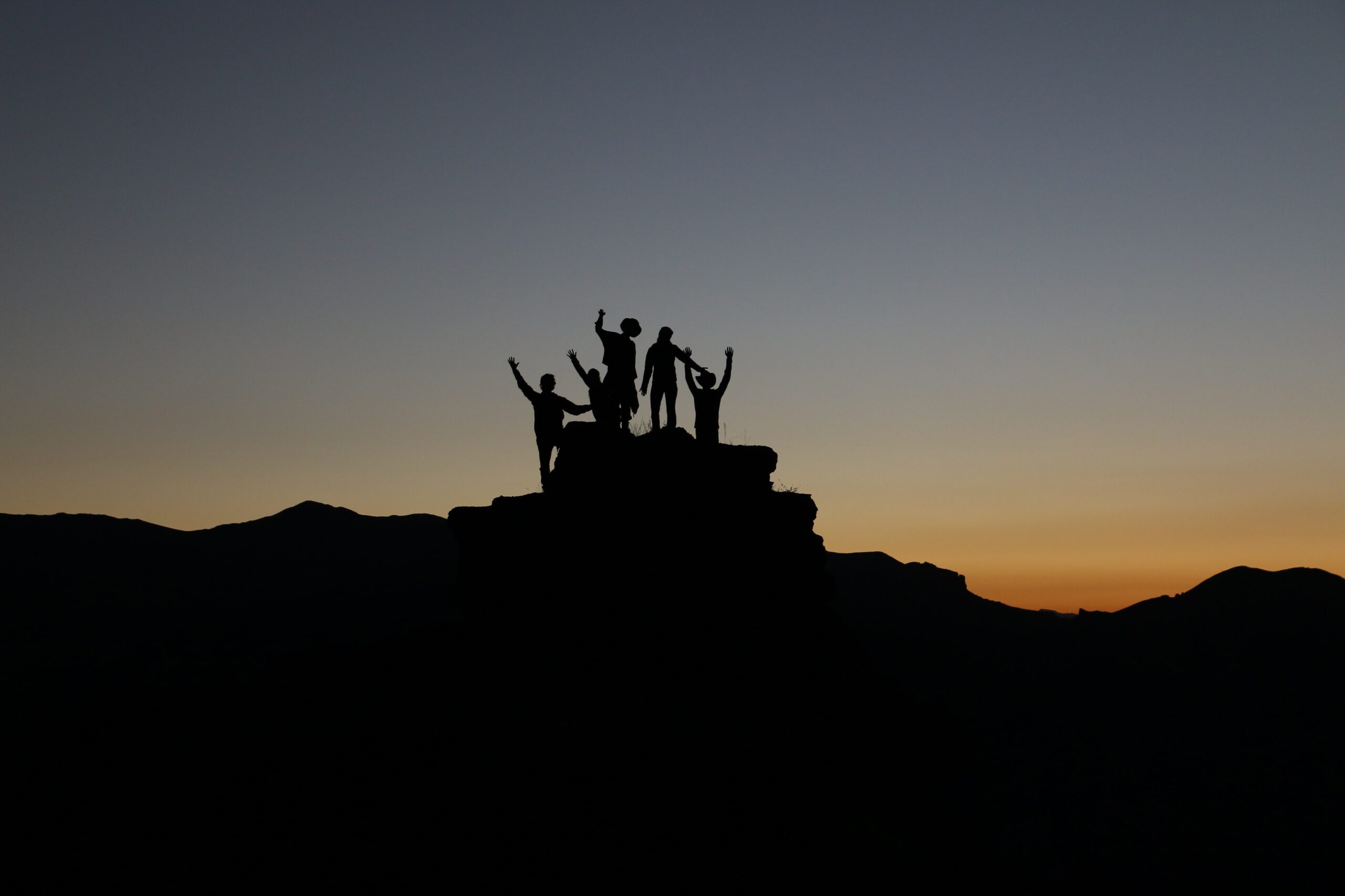 En gruppe mennesker står på toppen av et fjell ved solnedgang, med armene hevet i triumf. Silhuettene deres står mot en mørk himmel, og bildet formidler en følelse av prestasjon og fellesskap.