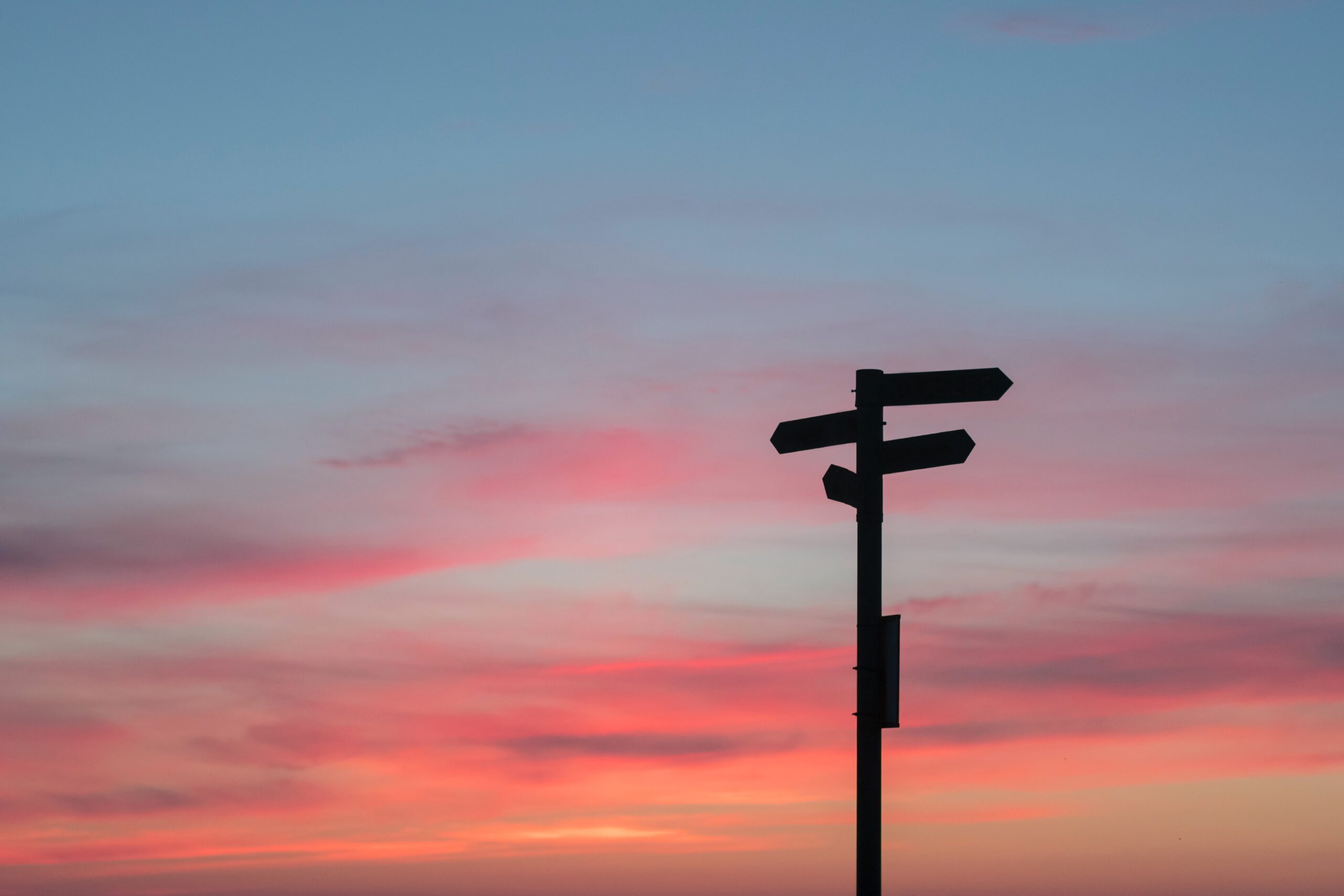 Et veiskilt med flere retninger mot en fargerik himmel ved solnedgang. Silhuetten av skiltet står tydelig mot den rosa og oransje bakgrunnen.