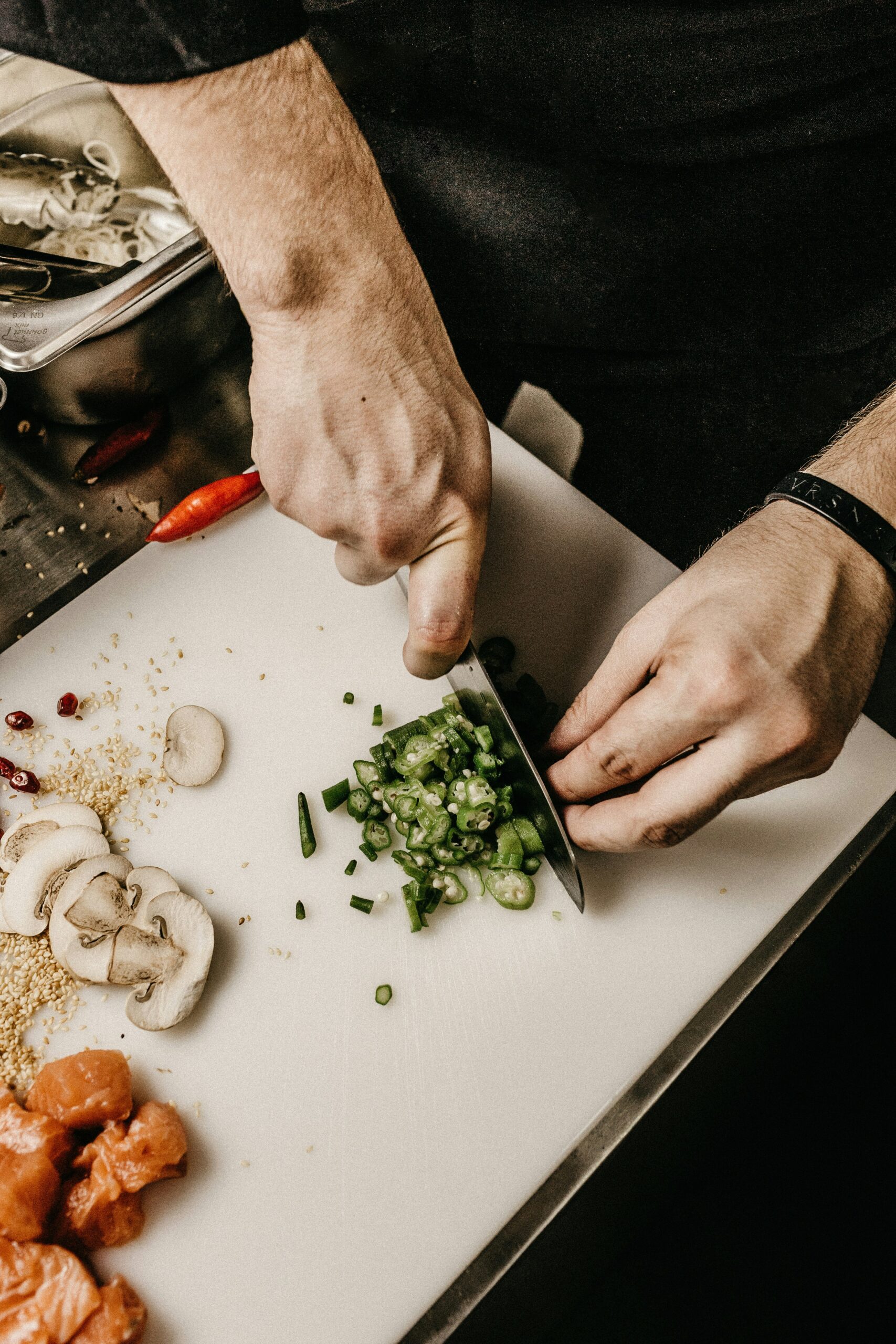 Nærbilde av hendene til en mann som hakker grønnsaker på et skjærebrett, omgitt av andre ingredienser som sopp og gulrøtter. Bildet gir et innblikk i matlagingsprosessen på kjøkkenet.