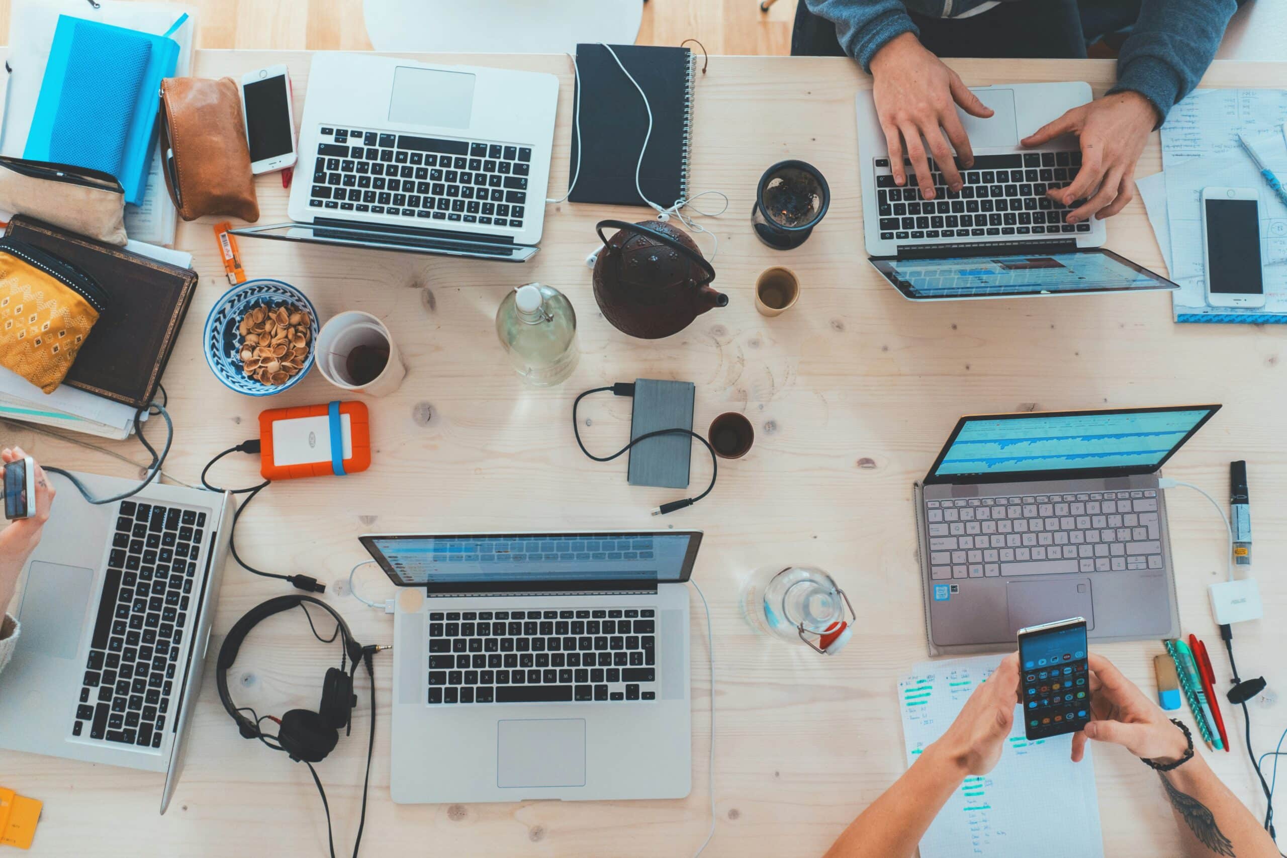Et fugleperspektiv av et bord med flere laptoper, kopper kaffe og kontorutstyr. Personer sitter rundt bordet og jobber, og scenen formidler en følelse av samarbeid og produktivitet.