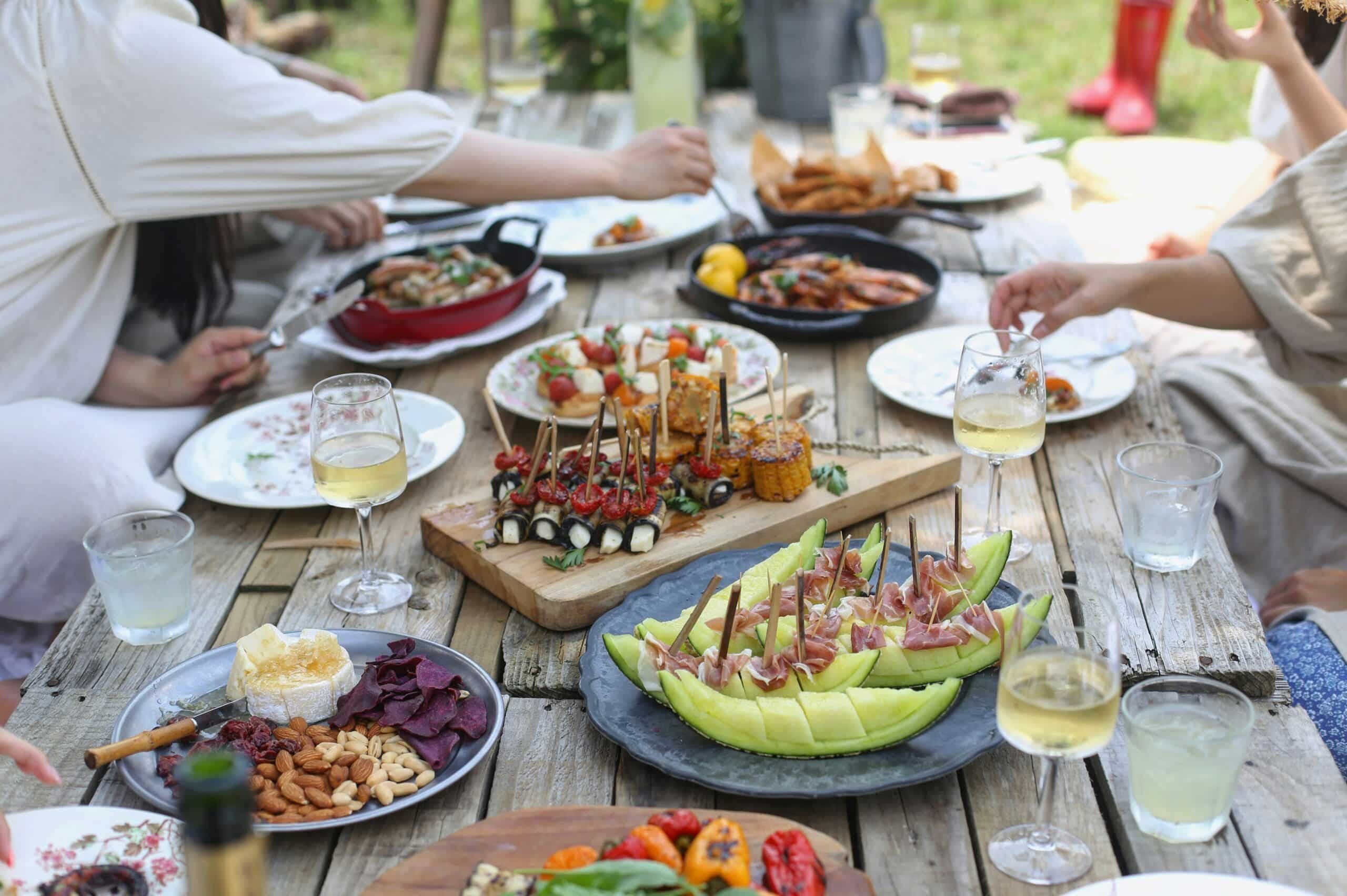 Et utendørs trebord dekket med ulike retter og tallerkener. Maten inkluderer ulike småretter som salater, grillmat, og frukt. Det er flere glass med hvitvin på bordet, og hendene til noen av gjestene er synlige i ferd med å forsyne seg. Bakgrunnen er grønn og gir en sommerlig atmosfære.