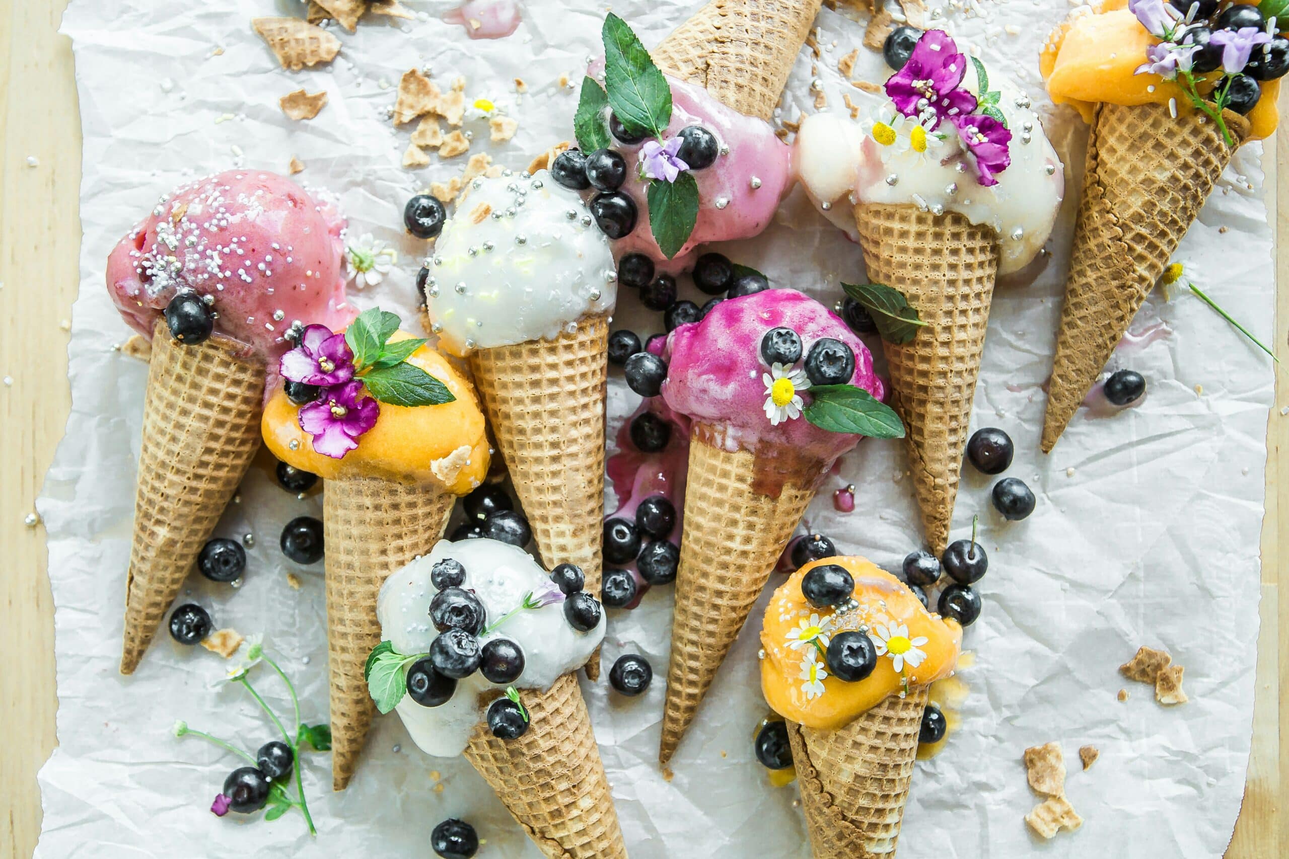 Flere iskjeks med fargerike iskuler ligger spredt på en lys overflate, pyntet med blåbær, spiselige blomster og myntegrener. Isen har ulike smaker og farger, inkludert rosa, hvit og oransje, som gir bildet et friskt og sommerlig uttrykk.