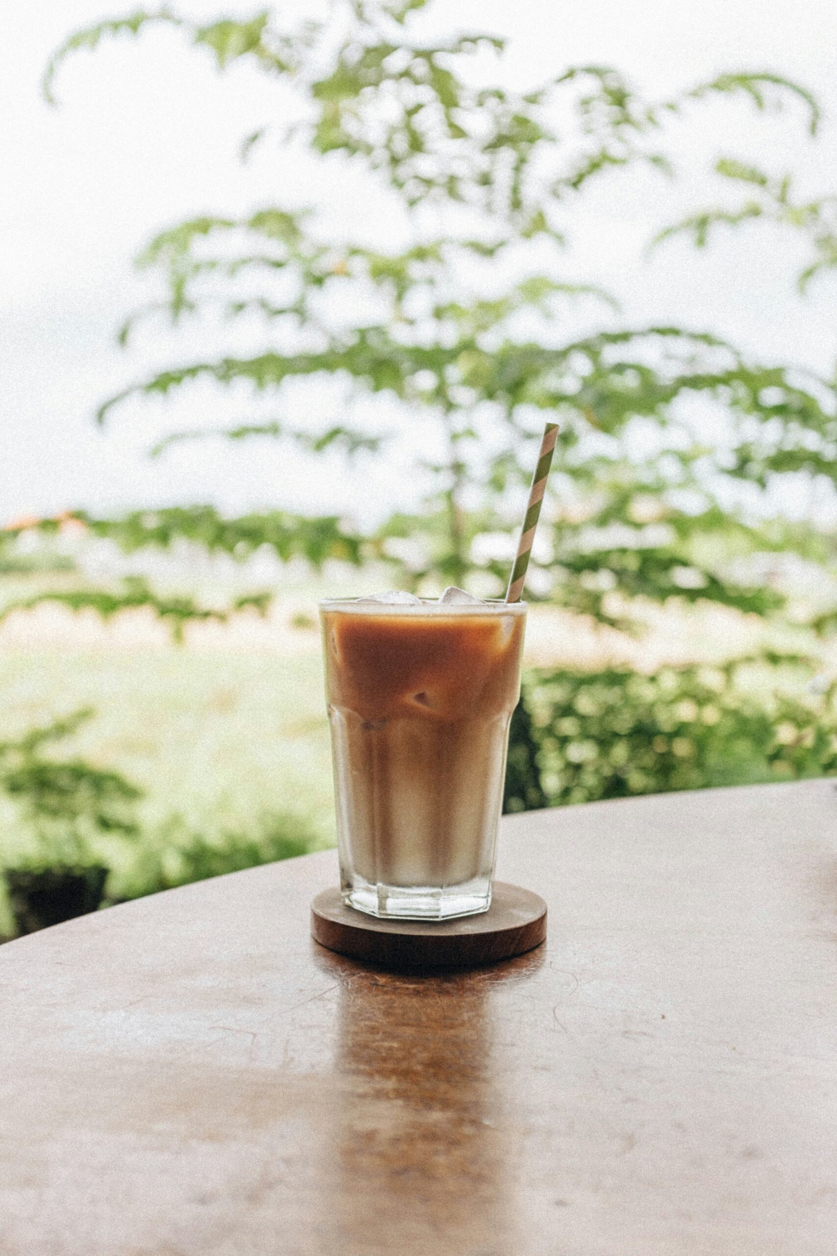 Et glass med iskaffe står på et rundt trebord utendørs. I bakgrunnen er det frodig grønn vegetasjon som skaper en avslappende og naturlig atmosfære. Iskaffen er lagdelt med melk og kaffe, og det står et metallisk sugerør i glasset.