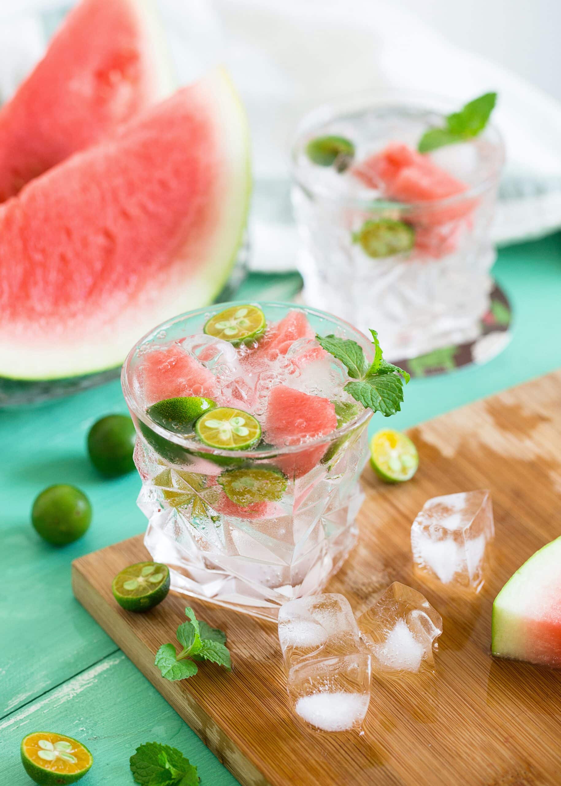 To glass fylt med isbiter, vannmelonbiter, lime og mynteblader står på et bord. I bakgrunnen er det en stor skive vannmelon. Scenen gir en frisk og sommerlig følelse, med lyse farger som dominerer bildet.