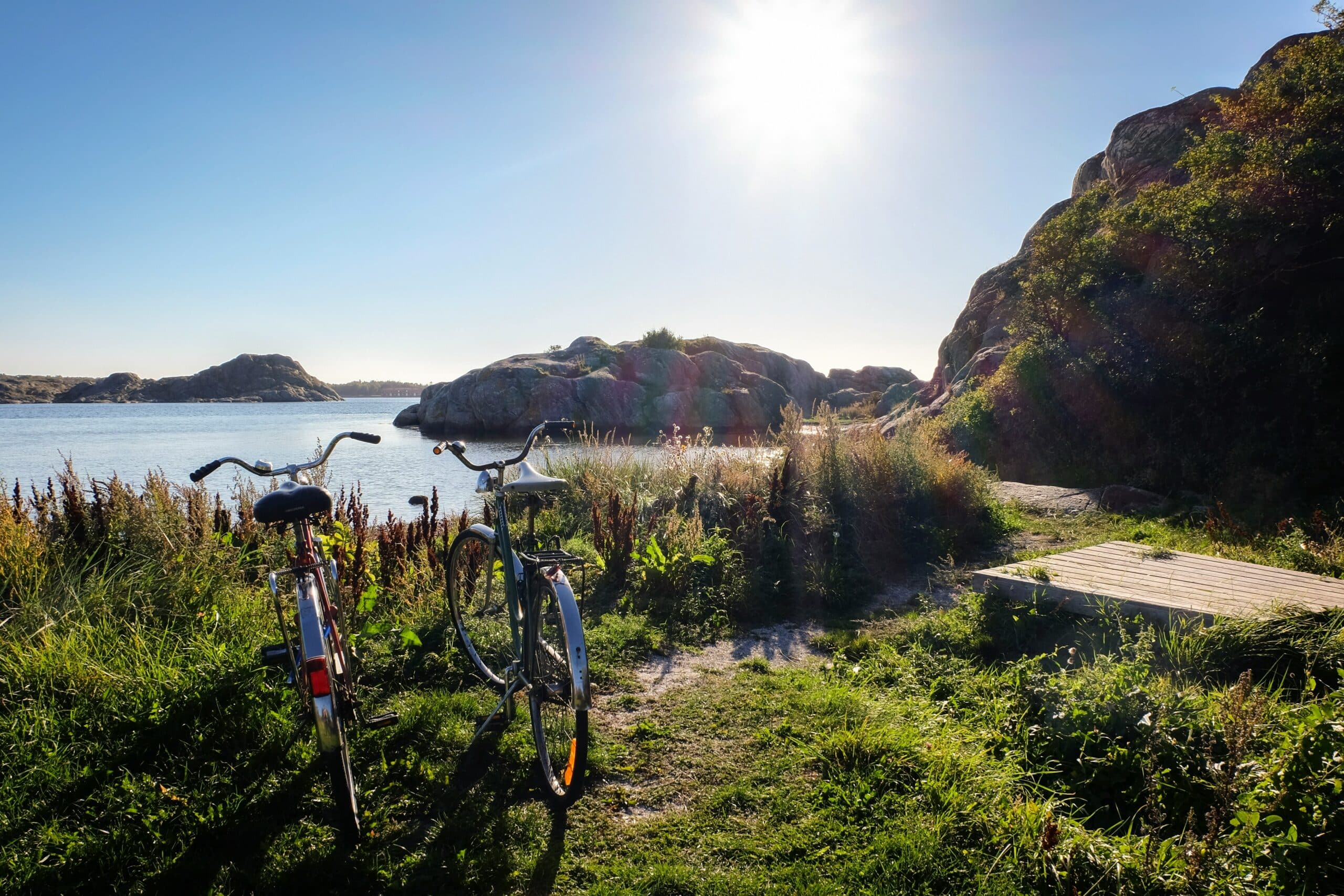 To sykler står parkert på en gresskledd sti ved sjøen, med utsikt mot et rolig vann og fjellformasjoner i bakgrunnen. Solen skinner klart på en blå himmel, og lyser opp det vakre kystlandskapet. Vegetasjonen rundt er grønn og frodig, og stien leder mot en treplatting på høyre side.