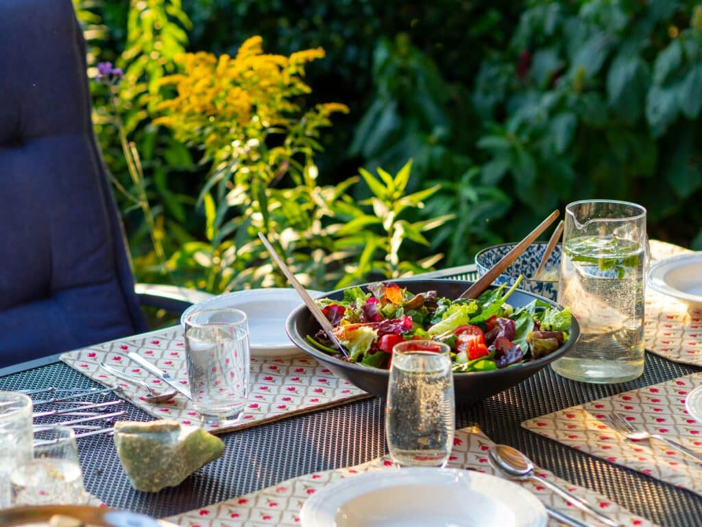 Et utendørs spisebord dekket med tallerkener, glass med vann, og en stor bolle med frisk salat. Bordet har dekorative bordbrikker, og i bakgrunnen er det frodig grønt løvverk som skaper en sommerlig og avslappet atmosfære. Sollyset skinner gjennom plantene og gir en varm og innbydende stemning.