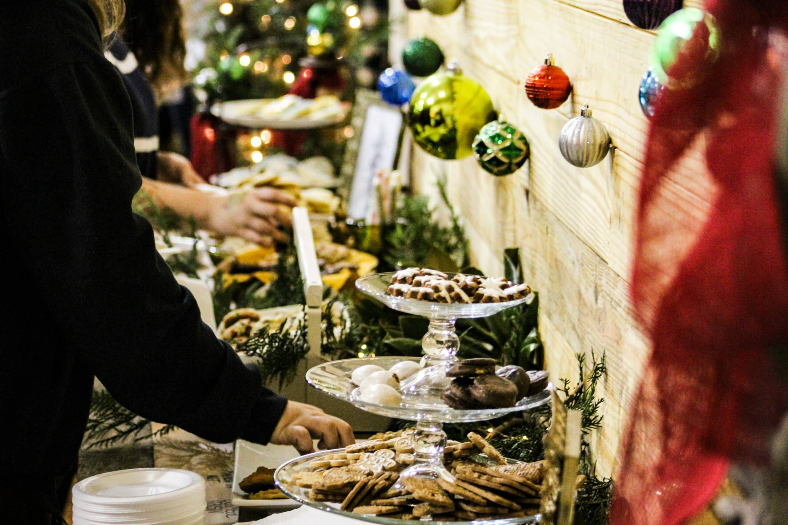 Et julebuffetbord med flere etasjefat fylt med forskjellige småkaker, inkludert pepperkaker og glasurdekkede kjeks. Bordet er pyntet med granbar og julepynt som fargerike julekuler. I bakgrunnen kan man skimte et juletre med lys, og folk som forsyner seg av maten.