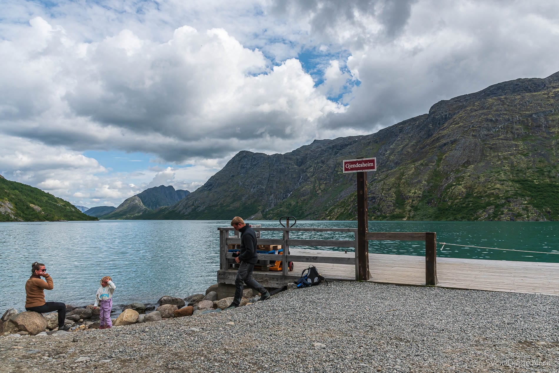 Bilde som viser tre personer som sitter ved kantet på Gjendevatnet. I bakgrunnen ser vi fjell.