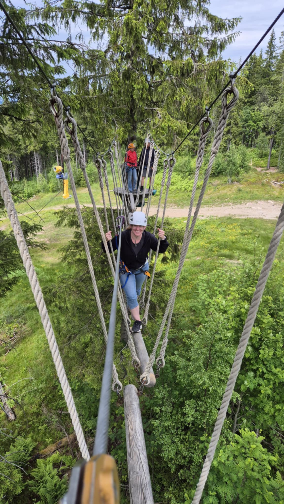 Klatrebane i tretopper, en kvinne med hjelm og klatresele balanserer i tauverk.