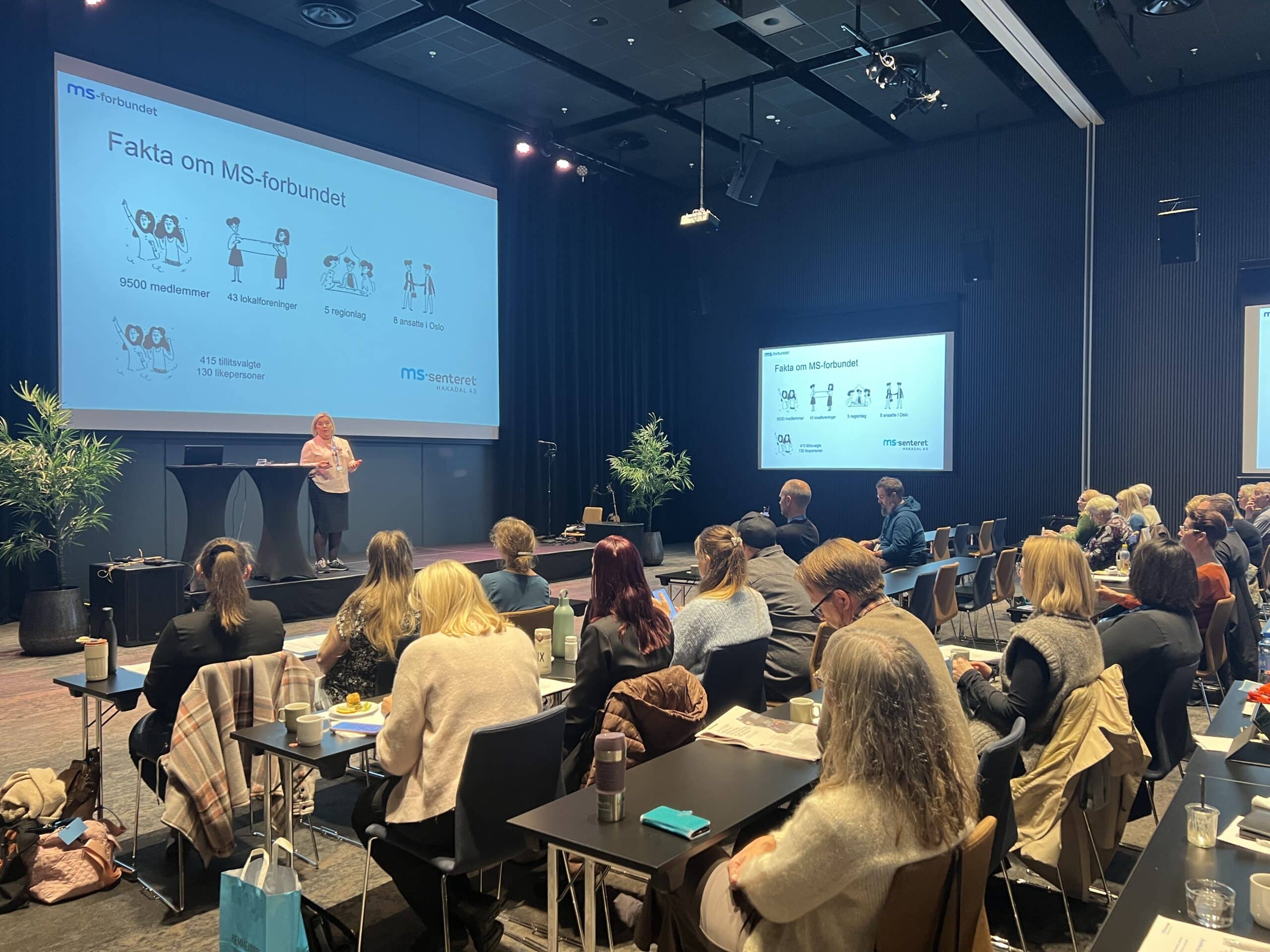 Bilde fra en MS-konferanse som viser en som står og holder presentasjon. Man ser bakhodene på publikum i salen.